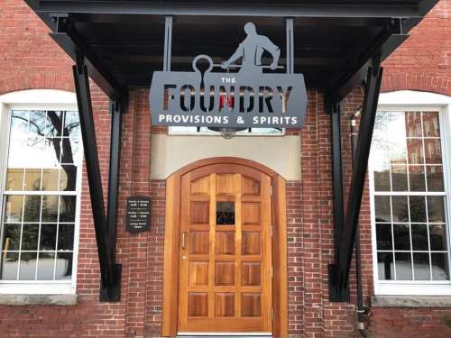 Sign for "The Foundry Provisions & Spirits" above a wooden door, set against a brick building with large windows.