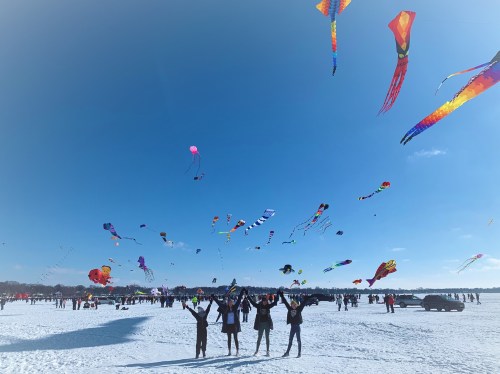A group of people stands on snow, joyfully raising their arms as colorful kites fly in a clear blue sky.