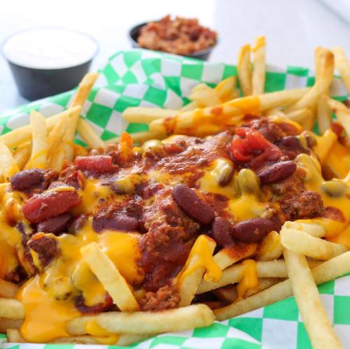 A basket of loaded fries topped with chili, cheese, and beans, served with dipping sauces in the background.