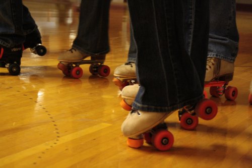 Close-up of roller skates on a wooden floor, with people wearing jeans and beige shoes.
