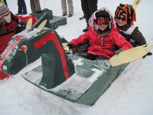 Two children in winter gear sit on a colorful, homemade sled shaped like a horse, surrounded by snow.