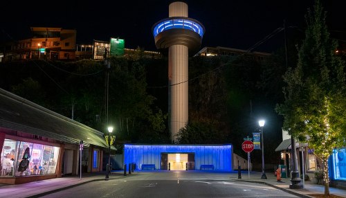 A tall tower with blue lighting stands over a quiet street, flanked by illuminated buildings at night.