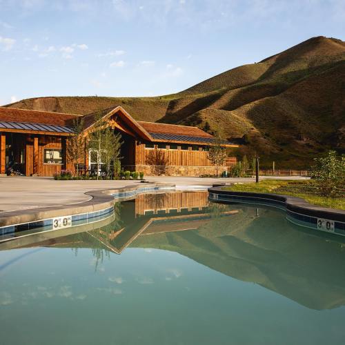 A serene pool area with wooden buildings and rolling green hills in the background under a clear blue sky.