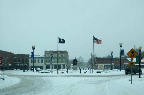 Snow-covered town square with flags flying, a statue in the center, and buildings partially obscured by falling snow.