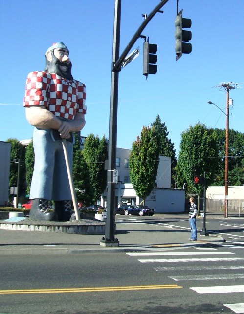 A giant statue of a lumberjack stands beside a traffic light, with a person walking nearby on a city street.