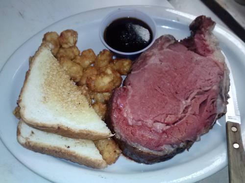 A plate featuring a large slice of prime rib, tater tots, two slices of bread, and a small cup of sauce.