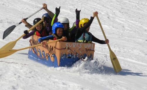 A group of six people in a colorful sled, paddling down a snowy slope, enjoying a fun winter activity.