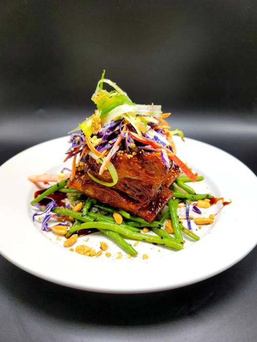 A plated dish featuring stacked glazed meat atop green beans, garnished with colorful vegetables and crushed peanuts.