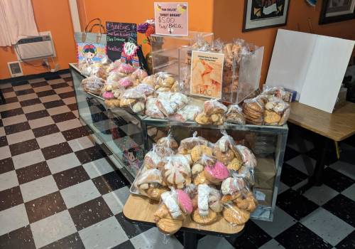 A display of assorted sweet breads in plastic bags on a table, with prices and colorful decorations in the background.