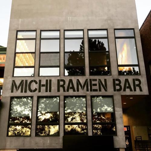 Exterior of Michi Ramen Bar, featuring large windows and a modern concrete facade. Reflections of trees are visible.