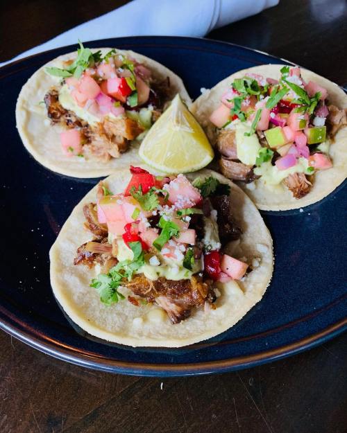 Three tacos on a plate, topped with diced vegetables, cilantro, and a lime wedge.