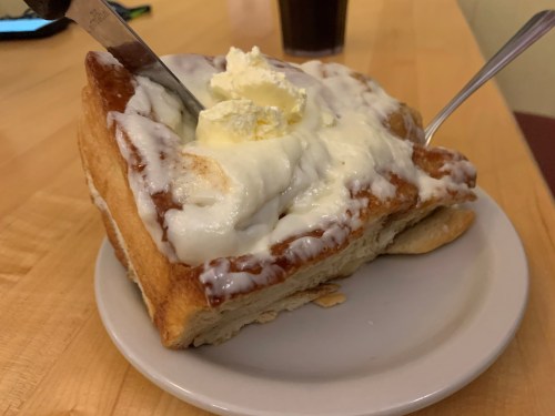 A large slice of cinnamon roll topped with cream cheese frosting and a pat of butter on a white plate.