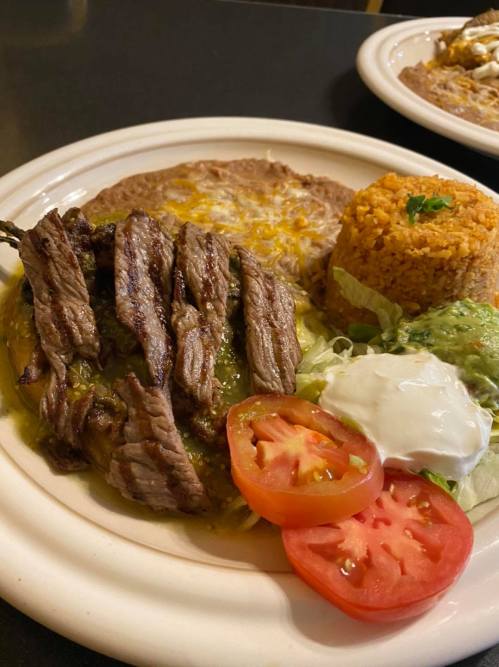A plate of grilled steak strips served with rice, beans, lettuce, tomato slices, and a dollop of sour cream.