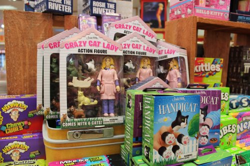 Three "Crazy Cat Lady" action figures with cats, surrounded by colorful candy boxes on a display table.