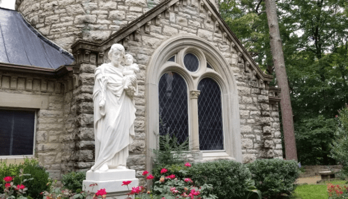 A stone building with a large window, featuring a white statue of a figure holding a child in front, surrounded by greenery.