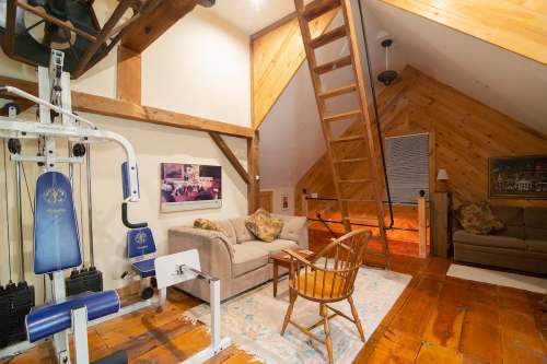 Cozy loft space with wooden beams, a workout machine, a sofa, and a ladder leading to an upper level.