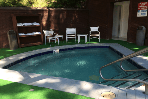 A small, inviting hot tub surrounded by green turf, with white chairs and a towel shelf nearby.