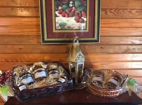 A wooden table displays two gift baskets with jars and a decorative lantern, set against a framed fruit painting.