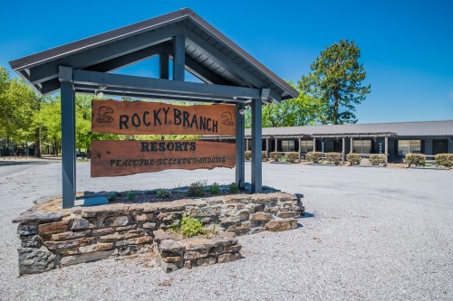 Sign for Rocky Branch Resorts, featuring a wooden design, surrounded by a gravel parking area and greenery.