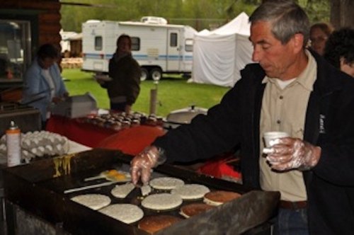 A man cooks pancakes on a griddle at an outdoor event, with people and tents in the background.