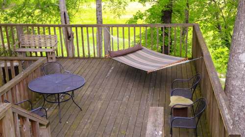 A wooden deck with a hammock, a round table, and chairs, surrounded by trees and greenery.