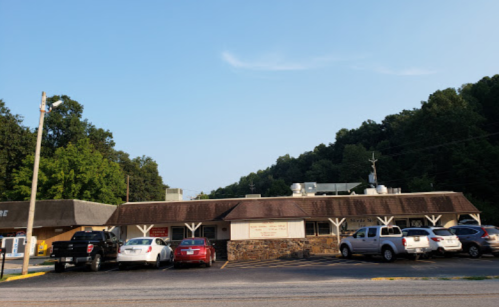 A small building with a stone facade and a sloped roof, surrounded by trees and parked cars.