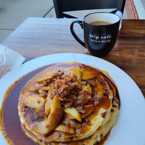 A plate of pancakes topped with apples and bacon, served with a cup of coffee on a wooden table.