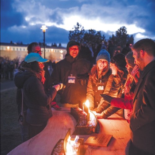 A group of people gathered around a fire pit, smiling and engaging in conversation during an outdoor event.