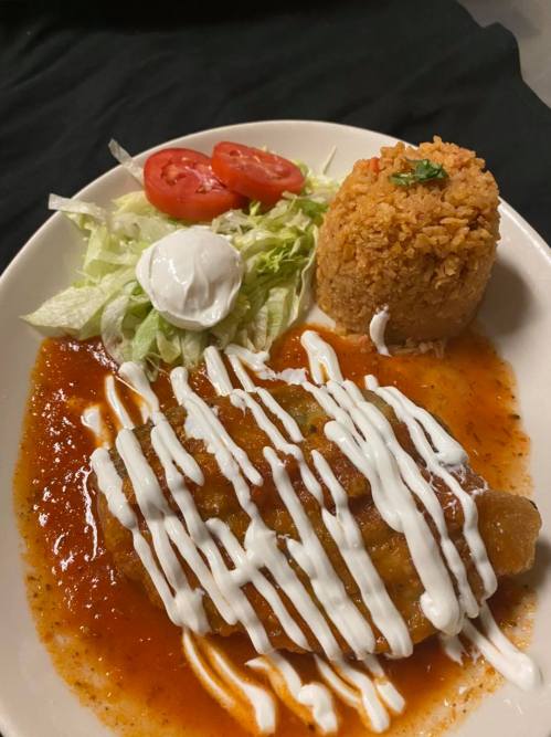 A plate of enchiladas topped with sauce and sour cream, served with rice, lettuce, and tomato slices.