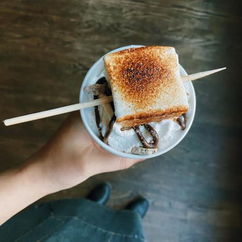 A hand holding a cup of dessert topped with toasted marshmallow and chocolate drizzle, resting on a wooden surface.
