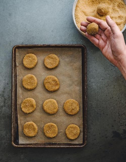 A hand holds a cookie while several rolled cookies coated in sugar sit on a baking tray lined with parchment paper.
