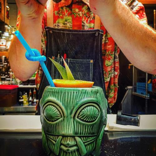 A colorful tiki cocktail in a green mug, garnished with an orange slice and leaves, with a bartender in a floral shirt.