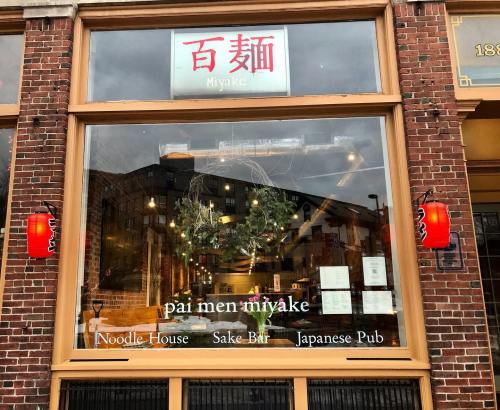 Window display of "Miyake," featuring signs for a noodle house, sake bar, and Japanese pub, with decorative lanterns.