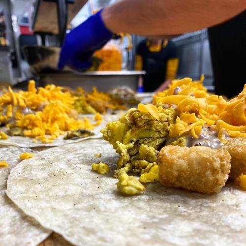A close-up of tortillas filled with scrambled eggs, tater tots, and shredded cheese, with a hand preparing food in the background.