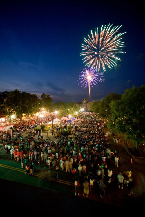A large crowd gathers in a park at night, watching colorful fireworks explode in the sky above.