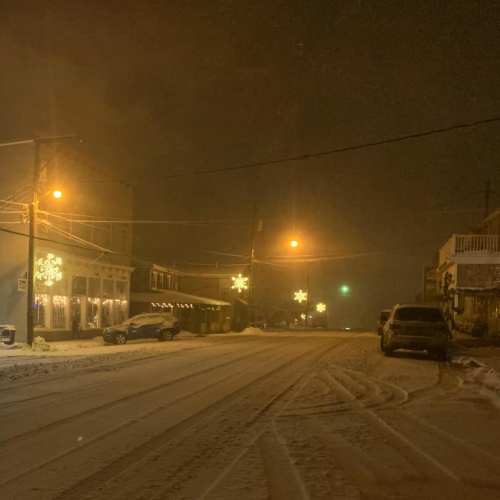 A snowy street at night, with festive lights and a few parked cars under a foggy sky.