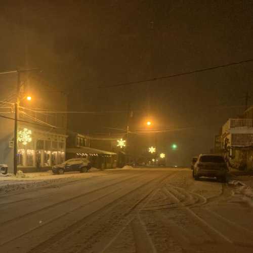 A snowy street at night, with festive lights and a few parked cars under a foggy sky.