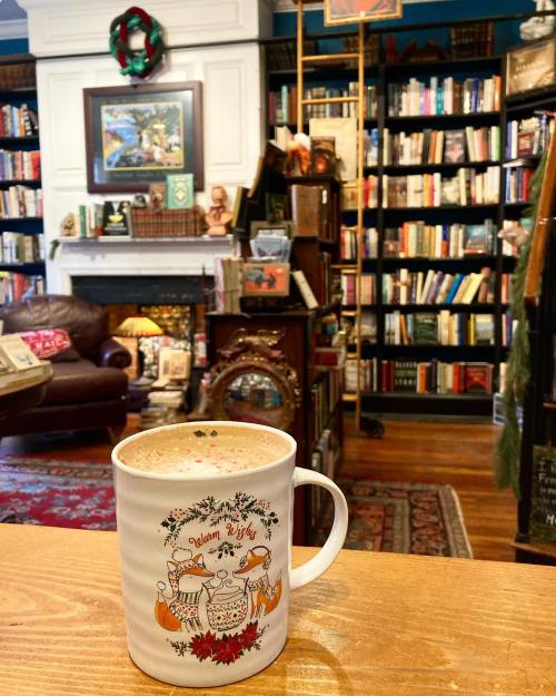 A cozy bookstore interior with a steaming mug in the foreground, surrounded by bookshelves and a warm atmosphere.