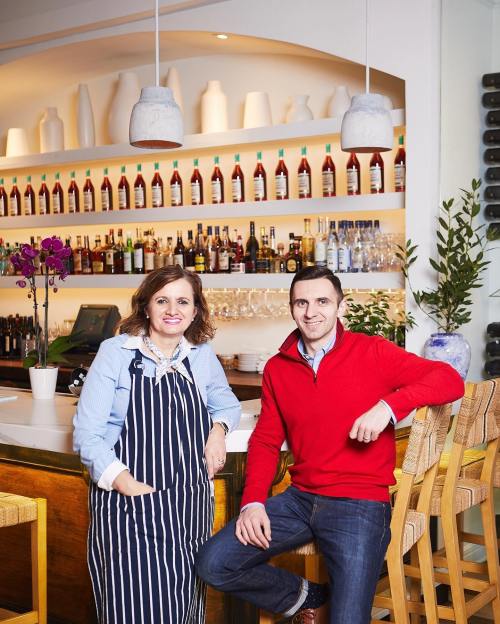 A woman in a striped apron and a man in a red sweater pose at a bar with shelves of bottles in the background.