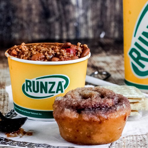 A bowl of chili with beans, a sweet cinnamon roll, and a Runza drink on a rustic wooden surface.