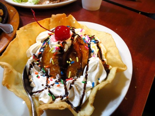 A dessert in a crispy bowl, featuring ice cream, whipped cream, chocolate sauce, sprinkles, and a cherry on top.