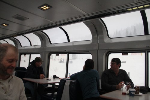 Interior of a train with passengers seated by large windows, overlooking a snowy landscape outside.