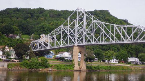 A steel bridge spans a river, with green hills and colorful houses visible on the opposite bank.