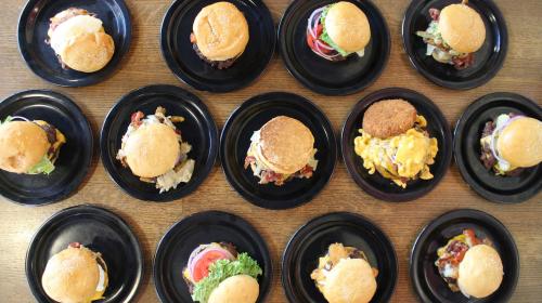A top-down view of several black plates, each with a burger and sides, arranged neatly on a wooden table.