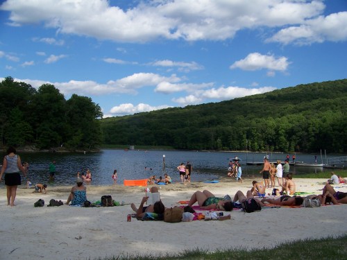 A sandy beach by a lake with people swimming, sunbathing, and enjoying the outdoors under a blue sky with clouds.