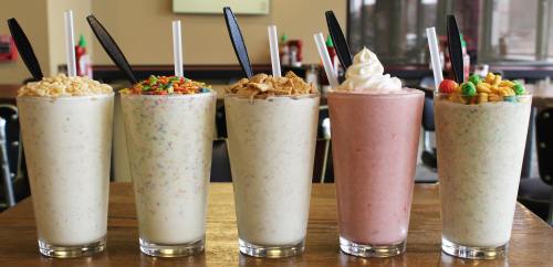 Five tall milkshakes in glasses, topped with colorful cereals and whipped cream, arranged on a wooden table.