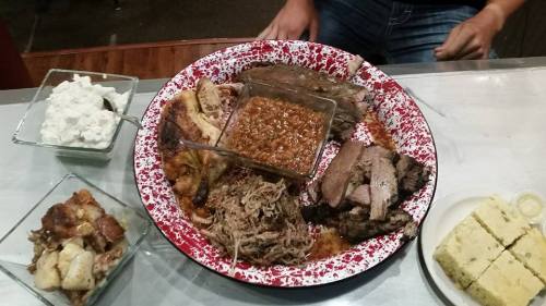 A large platter of assorted meats, baked beans, and sides, including cornbread and potato salad, on a rustic table.
