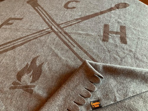 A gray blanket featuring a design with crossed matches, a flame, and the letters "FCH" in a rustic style.