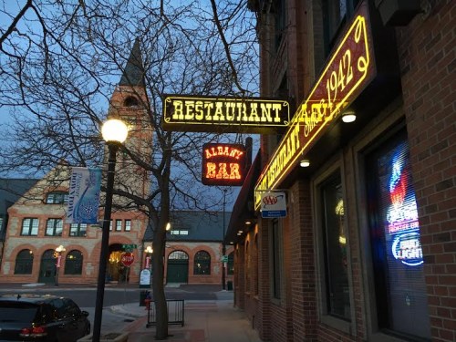 A street view of a restaurant sign and bar, with a historic building in the background during twilight.