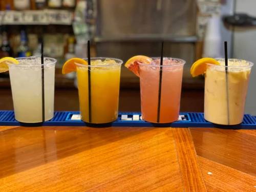 Four colorful cocktails with garnishes on a bar counter, featuring lemon and orange slices, served in clear cups.